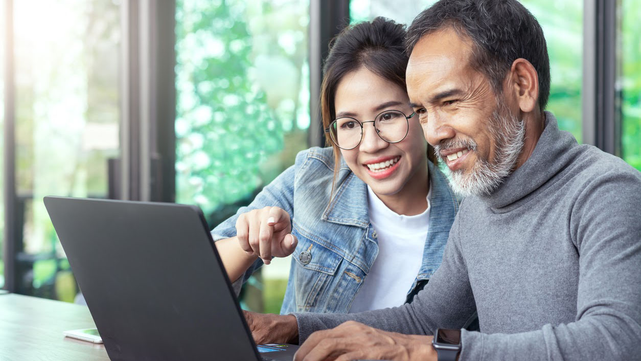 A photograph of a young woman helping an older man who is using a laptop